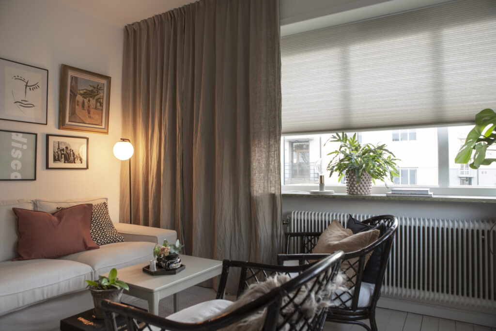 Beige duettegardin i gardinskenor bakom gardin i vardagsrum.